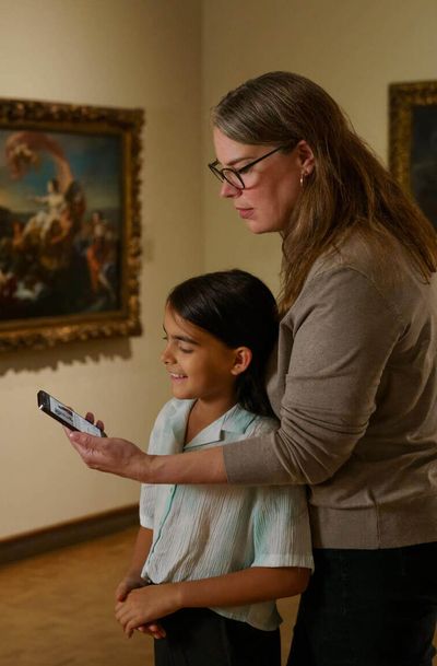 A parent holds their phone infront of a child, showing a guide to the European artwork they’re both standing infront of.
