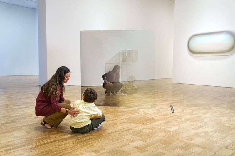 A parent squats down to discuss a contemporary art piece with their child who is seated on the floor of the museum.