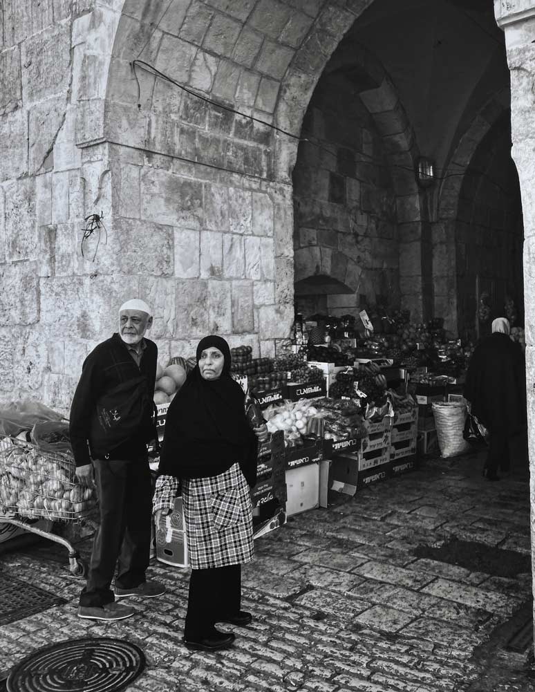 Elderly couple standing in an archway surrounded by marketplace goods