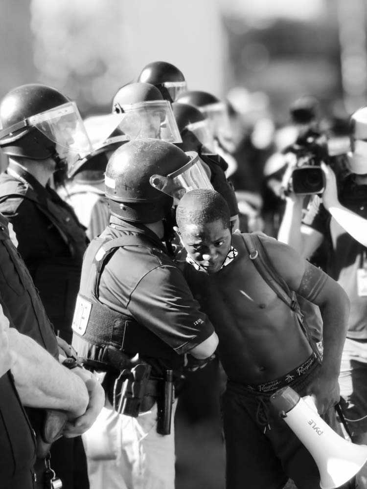 Young man holding a megaphone and locking arms with a police officer in riot gear surrounded by a line of other officers in gear
