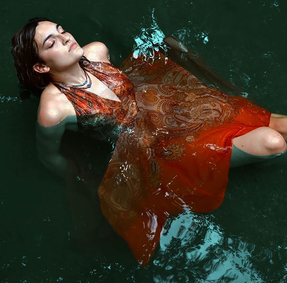 Young woman in a red paisley laying in shallow water with her head back and eyes closed