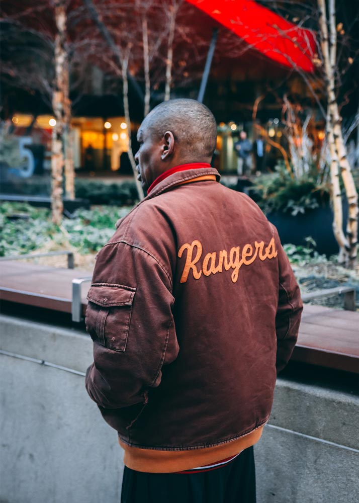 Man walking on the sidewalk wearing a jacket that says Rangers on the back