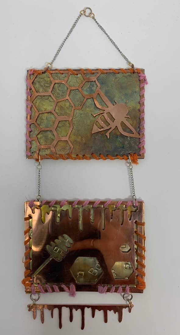 Two pieces of square metal connected together and hanging down, the top designed with a bee and honeycombs and the bottom with a stick of honey