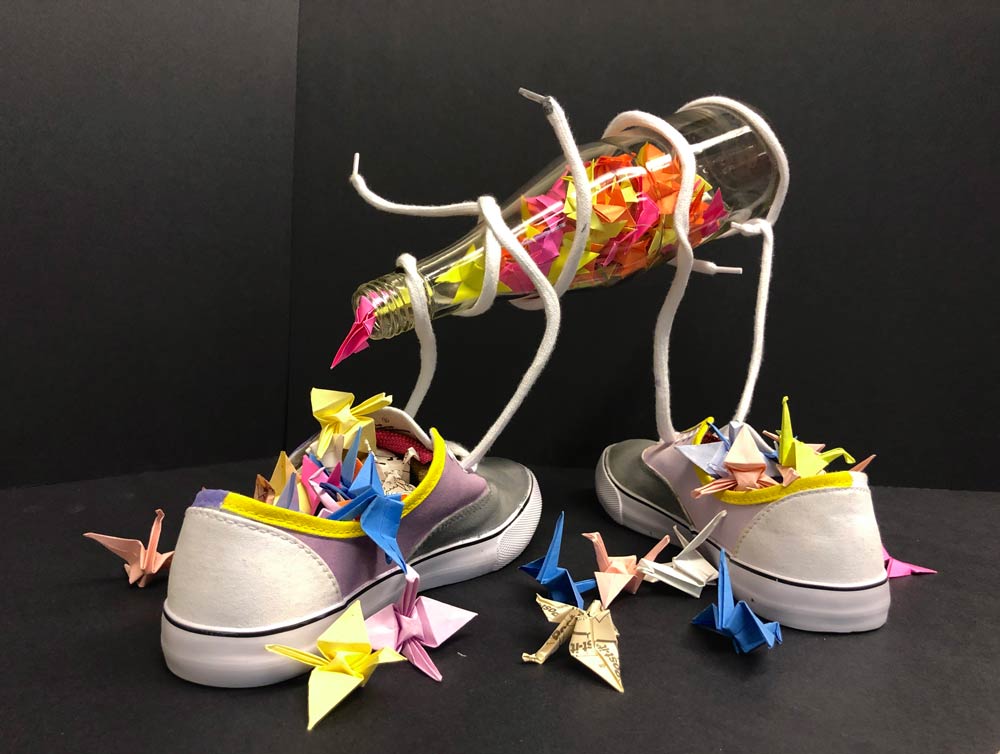 Pair of sneakers filled with colored pieces of construction paper with the shoestrings holding up a tilted glass soda bottle also filled with construction paper