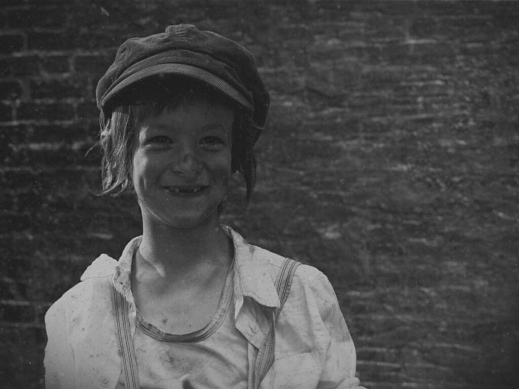 Black and white still of a young child in a cap and dirty clothing smiling for the camera