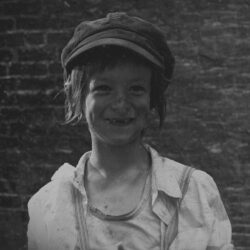 Black and white still of a young child in a cap and dirty clothing smiling for the camera