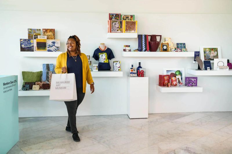 A museum guest holds a Milwaukee Art Museum Store shopping bag and smiles as they walk past a wall of collection artifacts available to purchase.