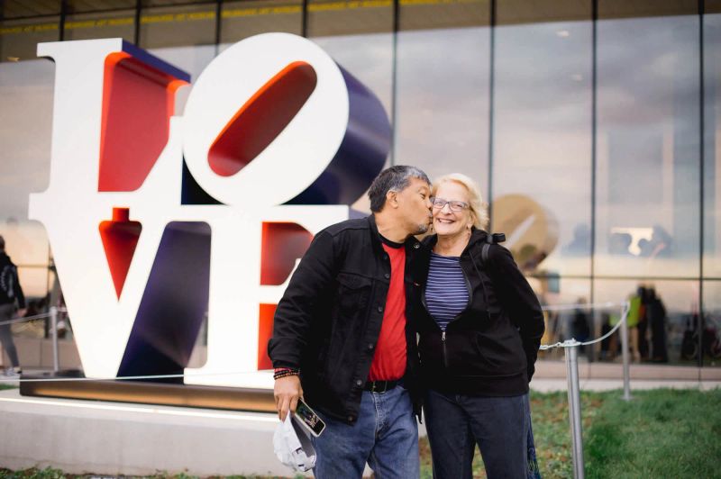 One museum guest kisses another museum guest on the cheek in front of Robert Indiana’s The American LOVE sculpture on the campus’s East End.