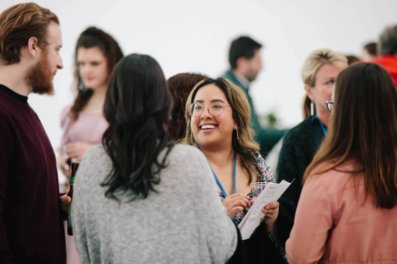 An event goer holds a drink and a paper agenda, and smiles at another guest who they’re in conversation with. They’re surrounded by other event goers.