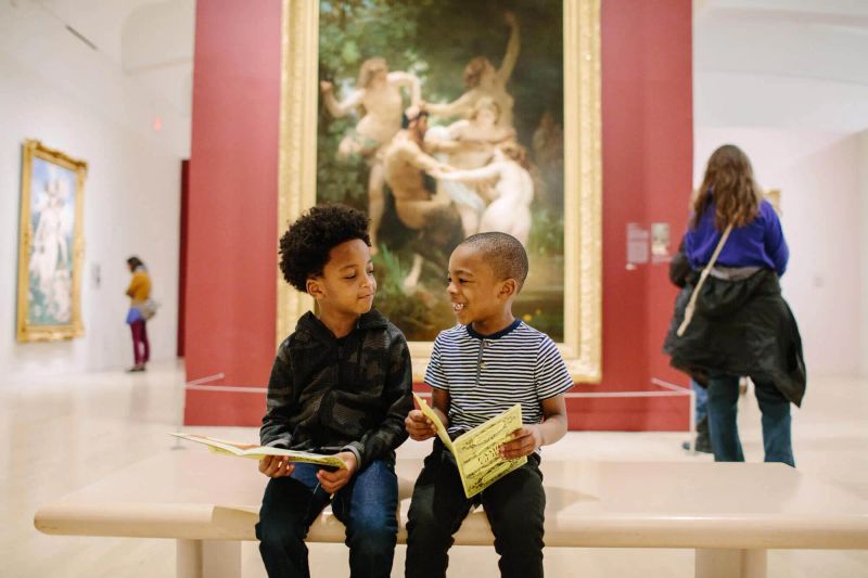 Two children sit in front of a large painting in the European collection, holding guides and smiling at each other.