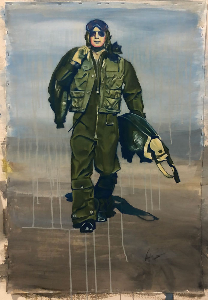 Man in a green one-piece flight suit with goggles on his head and a jacket over his shoulder