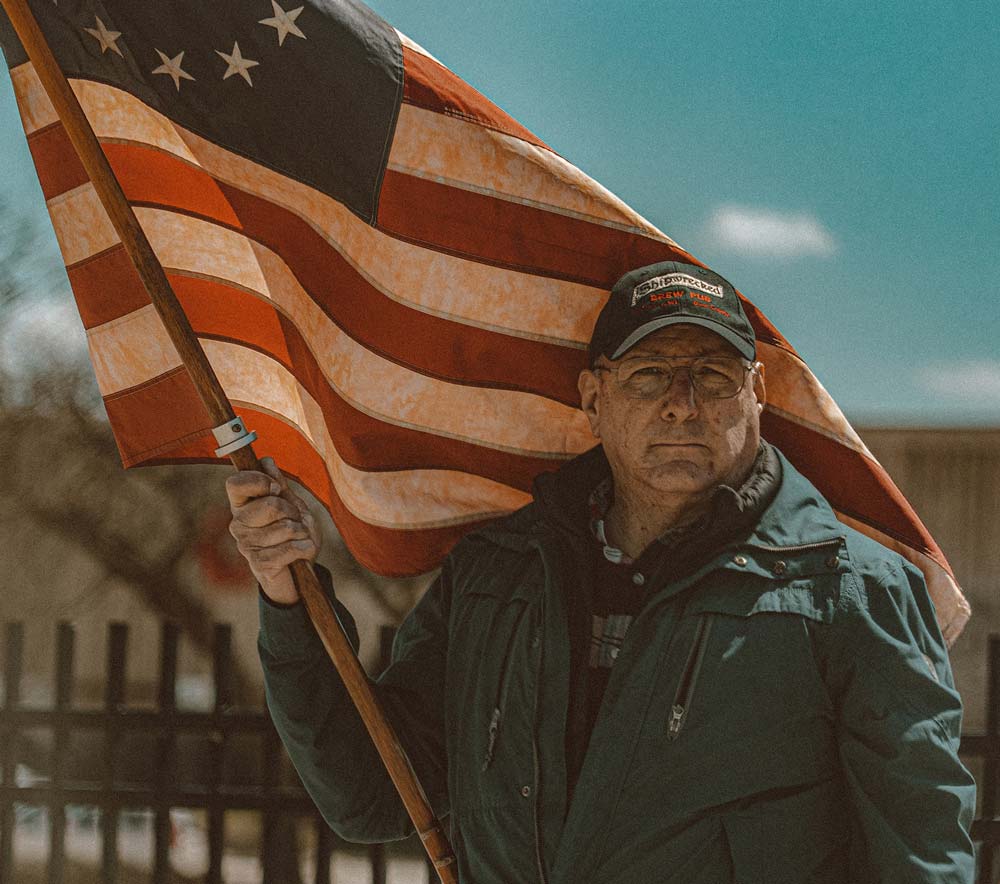 Older man in a thick jacket and baseball cap holding an American flag