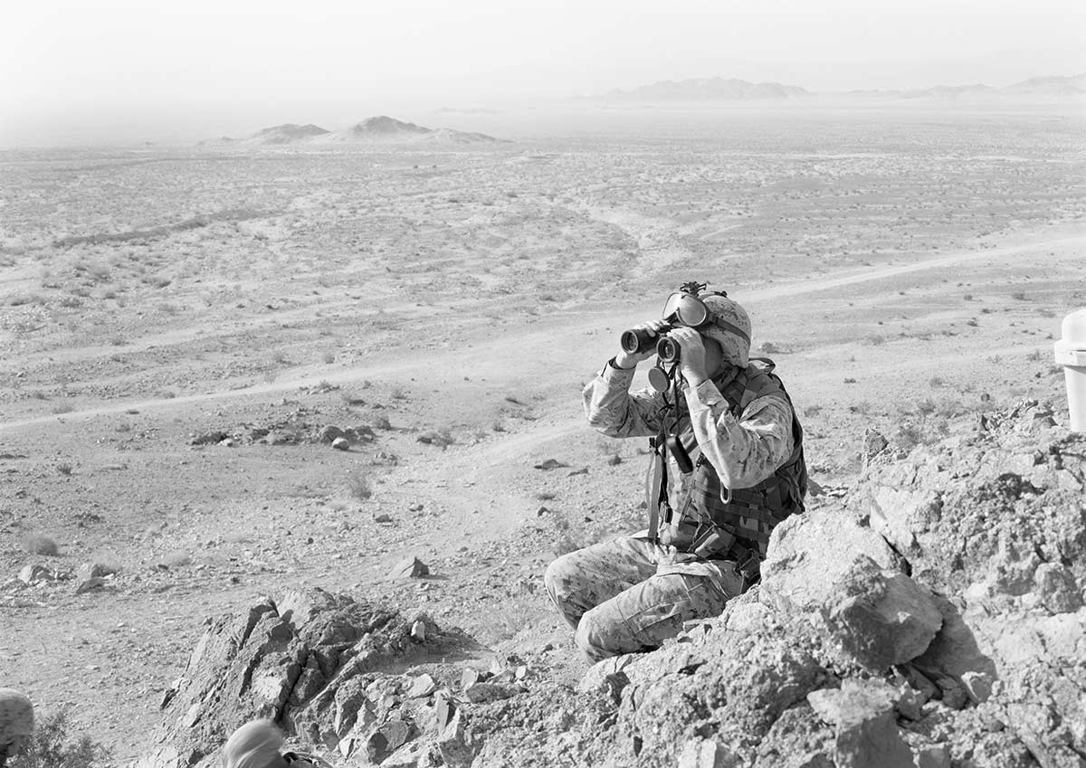 Soldier sitting in a desert looking through binoculars